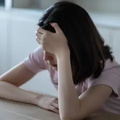 Kobieta z ręką na czole siedzi przy stole z kubkiem kawy.