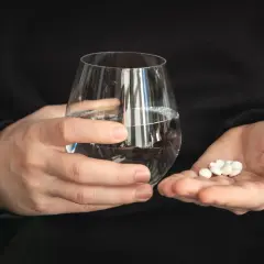 Dłonie trzymające szklankę wody i garść białych tabletek.