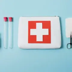 Apteczka z krzyżem, strzykawka, nożyczki, bandaże, probówki i leki na niebieskim tle.