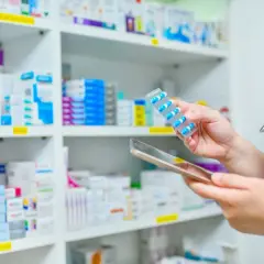 Farmaceuta z tabletką w ręku i stetoskopem na szyi, na tle półek z lekami.