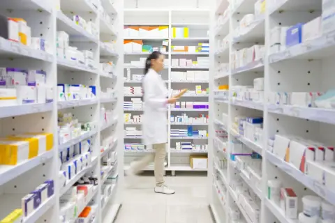 Farmaceutka w białej kitce porusza się między regałami pełnymi leków, szukając potrzebnego produktu. W aptece, gdzie panuje porządek, można znaleźć wiele preparatów, w tym te z grupy polpril.