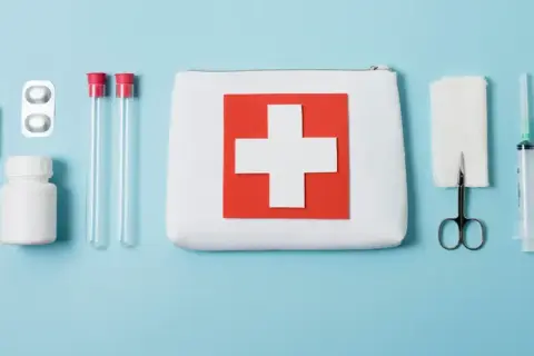 Apteczka z krzyżem, strzykawka, nożyczki, bandaże, probówki i leki na niebieskim tle.