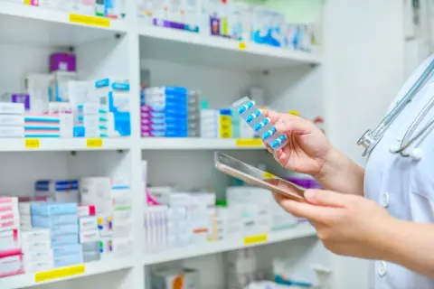 Farmaceuta z tabletką w ręku i stetoskopem na szyi, na tle półek z lekami.