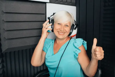 Uśmiechnięta kobieta w słuchawkach podczas badania audiometrii tonalnej. Pokazuje kciuk w górę, co oznacza pozytywny wynik.